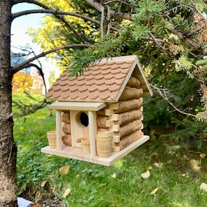 May include: A wooden birdhouse shaped like a cabin with a cork roof and cork siding. The birdhouse has a small opening for birds to enter and a small wooden porch with two small wooden barrels.