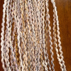 May include: A close-up of a bundle of yarn in shades of beige, brown, and white with a subtle shimmer. The yarn is twisted and textured, creating a unique and interesting look.