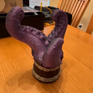 May include: A purple tentacle sculpture with a textured surface and suction cups. The base is a dark brown barrel shape with grey spikes. The sculpture is on a wooden table.