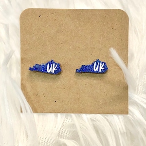 May include: A pair of blue glitter Kentucky-shaped stud earrings. Each earring has the letters "UK" in white. The earrings are displayed on a brown card against a white, fluffy background.
