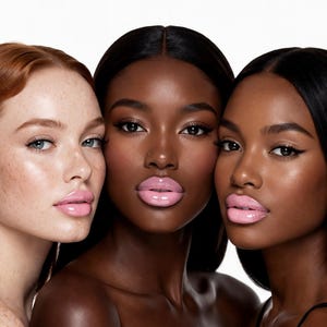 May include: Three women with different skin tones pose for a beauty shot. The woman on the left has red hair and freckles, the woman in the middle has dark skin, and the woman on the right has dark skin. All three women have glossy pink lipstick.