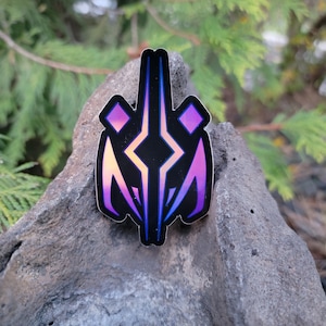 May include: A holographic sticker with a purple, blue, and pink geometric design. The sticker features a stylized, abstract shape with sharp angles and a diamond-like center.