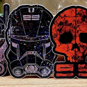 May include: Set of six black and white stickers featuring Star Wars clone trooper helmets with red and blue accents. The helmets have a distressed, glittery look. One sticker features a red skull with a cracked surface.