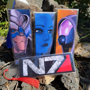May include: Four clear plastic bookmarks with colorful images and tassels. The bookmarks feature characters from the video game Mass Effect. One bookmark has a blue and white image of a character with a helmet. Another bookmark has a blue image of a female character with blue eyes. The third bookmark has a purple and blue image of a character with a helmet. The fourth bookmark has a black and white image of the Mass Effect logo with the text "N7".