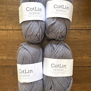 Può includere: Quattro gomitoli di filato grigio con etichette bianche che riportano "CotLin DK WEIGHT". Ogni gomitolo è etichettato come 123 iarde/50 grammi. Il filato è disposto su una superficie di legno.