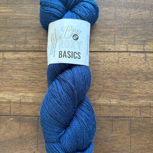 May include: A skein of deep blue yarn with a white label that reads "Leo & Roxy Basics". The yarn is tightly wound and appears soft. The background is a wooden surface.
