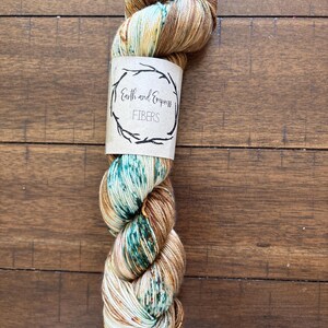 May include: A skein of hand-dyed yarn in shades of brown, teal, and cream, with a label that reads "Earth and Empress FIBERS." The yarn is twisted and displayed on a wooden surface, showcasing its color variations.