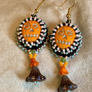 May include: A pair of orange and black beaded earrings with a skull design and a bell charm. The earrings are made with small beads and have a gold hook.