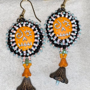 May include: A pair of orange and black beaded earrings with a skull design and a bell charm. The earrings are made with black, white, and green beads.