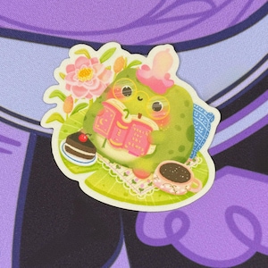 May include: A whimsical sticker featuring a green frog wearing glasses and a pink hat, reading a book titled "Year for the Stars." The frog is surrounded by a slice of cake, a cup of coffee, and flowers. The sticker has a white border.