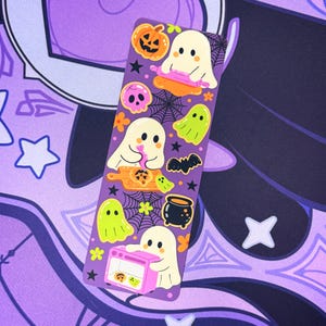 May include: A purple bookmark featuring Halloween-themed illustrations. The design includes cartoon ghosts, pumpkins, a skull, a cauldron, a bat, and spiderwebs. The ghosts are depicted in various activities, such as baking and eating. The bookmark is decorated with yellow and orange accents.