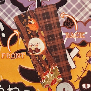 May include: A double-sided bookmark with a woodland scene on the front and a brown and white plaid pattern on the back. The front features a white owl with a book, mushrooms, and leaves in a forest setting.