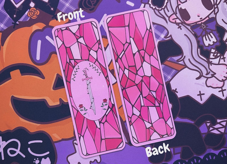 May include: Two pink rectangular tarot cards. The front card features a sword design with roses and a wreath, while the back has a stained glass pattern. The cards are set against a purple background with a jack-o'-lantern and Japanese text.