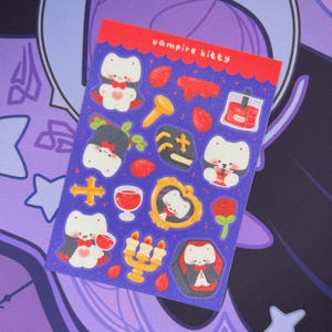 May include: A sticker sheet with a purple background, featuring various vampire-themed illustrations. The stickers depict a cartoon cat in a vampire costume, crosses, and other spooky elements. The text "vampire kitty" is printed at the top.