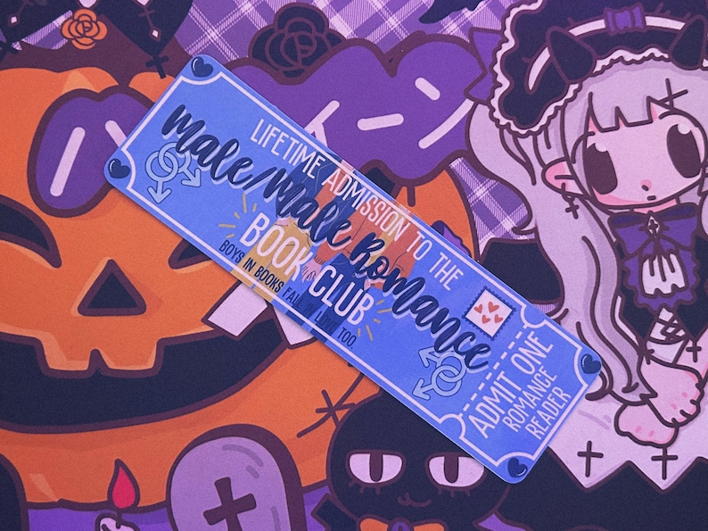 May include: A blue bookmark with the text "Male/Male Romance Book Club" and "Lifetime Admission". The bookmark has a ticket design with hearts and male gender symbols. The background features Halloween-themed illustrations.