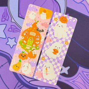 May include: Two decorative bookmarks with Halloween-themed designs. One features a pumpkin cottage, ghosts, and pumpkins on a pink background. The other has ghosts and pumpkins on a checkered purple and white background. Both have a whimsical, cartoonish style.