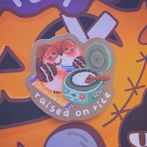 May include: A sticker featuring two cartoon characters with rice bowls, and a rice cooker with the text "raised on rice". The sticker has a brown border and is set against a patterned orange background.