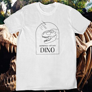 以下が含まれることがあります： 恐竜の頭部の黒い線画と「Definitely not our Dino」というテキストが書かれた白いTシャツ。