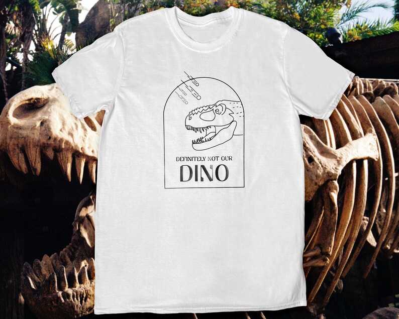 以下が含まれることがあります： 恐竜の頭部の黒い線画と「Definitely not our Dino」というテキストが書かれた白いTシャツ。