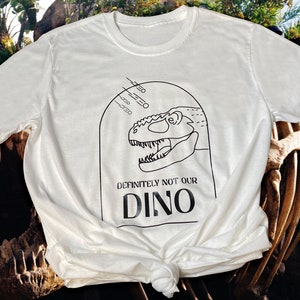 以下が含まれることがあります： 黒い恐竜の頭と「Definitely not our Dino」というテキストが書かれた白いTシャツ