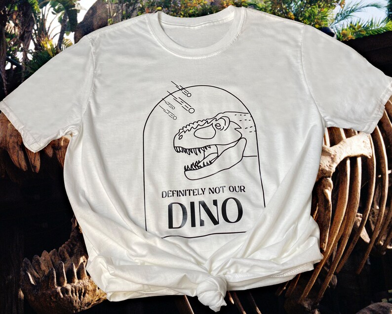 以下が含まれることがあります： 黒い恐竜の頭と「Definitely not our Dino」というテキストが書かれた白いTシャツ