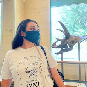 以下が含まれることがあります： 白のTシャツを着た女性。Tシャツには、黒と白の恐竜のグラフィックと「DEFINITELY NOT OUR DINO」という文字がプリントされています。彼女は緑色のマスクを着けており、背景の恐竜の頭蓋骨を見ています。