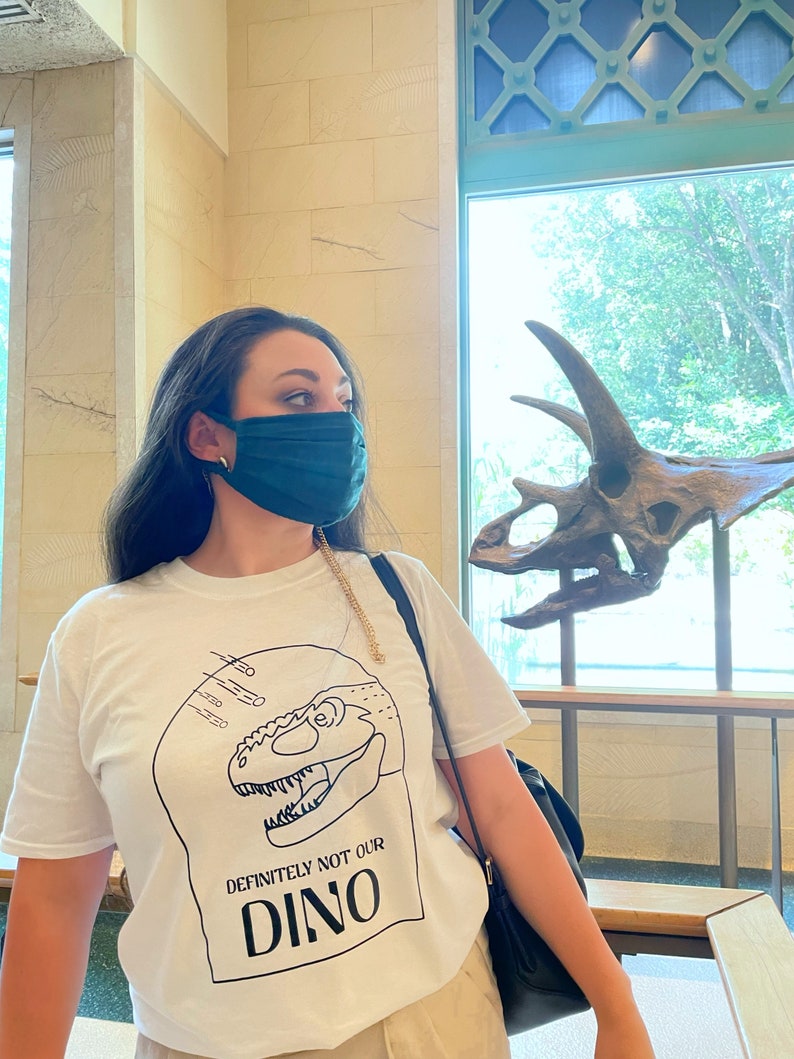 以下が含まれることがあります： 白のTシャツを着た女性。Tシャツには、黒と白の恐竜のグラフィックと「DEFINITELY NOT OUR DINO」という文字がプリントされています。彼女は緑色のマスクを着けており、背景の恐竜の頭蓋骨を見ています。