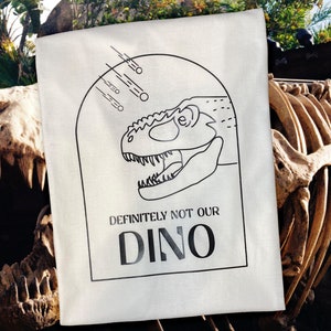 以下が含まれることがあります： 白いトートバッグに、黒い線で描かれた恐竜の頭と「DEFINITELY NOT OUR DINO」という文字が書かれています。