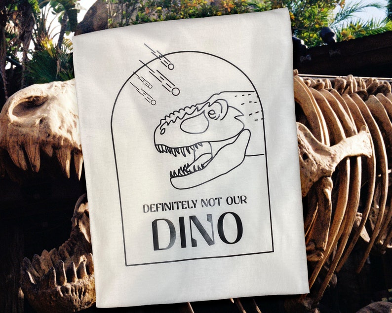 以下が含まれることがあります： 白いトートバッグに、黒い線で描かれた恐竜の頭と「DEFINITELY NOT OUR DINO」という文字が書かれています。