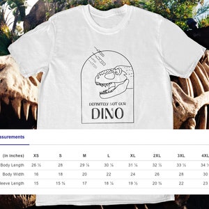 以下が含まれることがあります： 白いTシャツに、黒い線で描かれた恐竜の頭と「DEFINITELY NOT OUR DINO」という文字が書かれています。Tシャツには、インチで測定されたサイズ表が付いています。