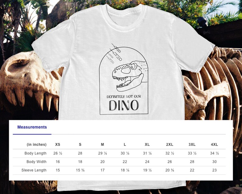 以下が含まれることがあります： 白いTシャツに、黒い線で描かれた恐竜の頭と「DEFINITELY NOT OUR DINO」という文字が書かれています。Tシャツには、インチで測定されたサイズ表が付いています。