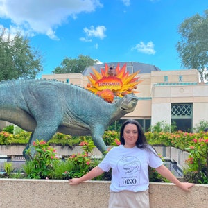 以下が含まれることがあります： 「Definitely not for Dino」という黒い文字と恐竜のグラフィックが書かれた白いTシャツを着た人がいます。その人は、「Dinosaur」という赤い文字が書かれた看板のある大きな恐竜の像の前に立っています。