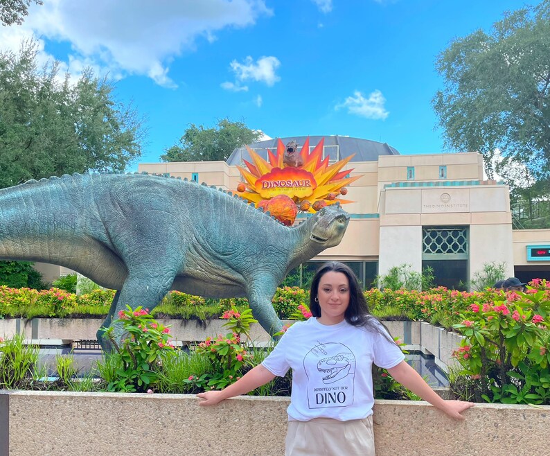 以下が含まれることがあります： 「Definitely not for Dino」という黒い文字と恐竜のグラフィックが書かれた白いTシャツを着た人がいます。その人は、「Dinosaur」という赤い文字が書かれた看板のある大きな恐竜の像の前に立っています。