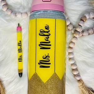 May include: A yellow and gold glitter tumbler with a black pencil design and the text "Mrs. Mallo" on it. The tumbler has a pink lid with a white "Lift to Unlock" label.