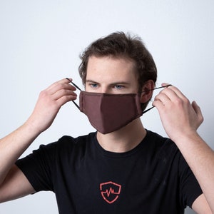 May include: A person wearing a brown face mask with black elastic ear loops. The mask is made of a soft, breathable fabric.