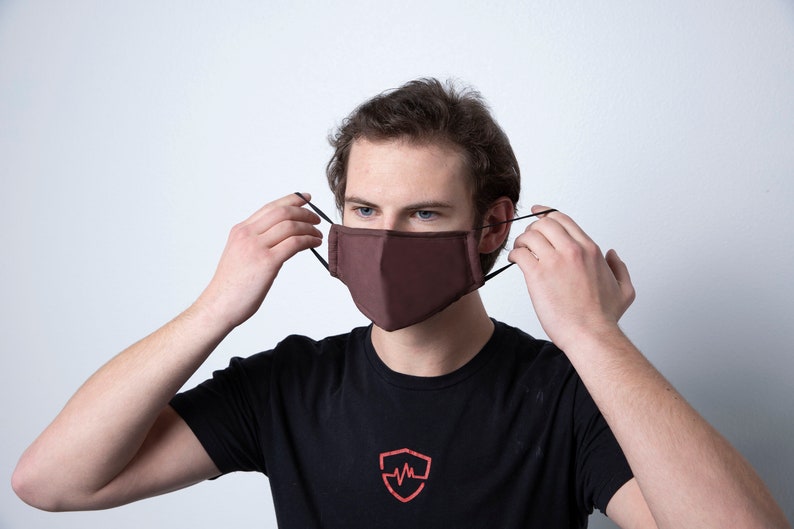 May include: A person wearing a brown face mask with black elastic ear loops. The mask is made of a soft, breathable fabric.