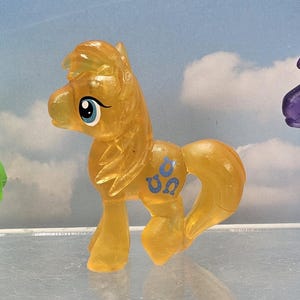 May include: Three small pony figurines made of translucent plastic. The left pony is bright green with a flower symbol. The center pony is amber yellow with a blue horseshoe symbol. The right pony is deep purple with a white musical note symbol.