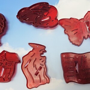 May include: Six red plastic cookie cutters in various shapes, including a heart, a rabbit, a butterfly, a boot, a witch, and a flag. The cutters are arranged on a light blue background.