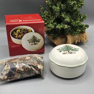 May include: A white ceramic Christmas tree potpourri jar with a green lid and a green and red Christmas tree design. The jar is filled with brown, green, and red potpourri. The jar is sitting on a brown surface next to a box with the words "Christmas Morning" printed on it.