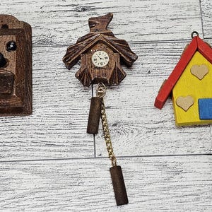 May include: Three wooden decorative items: a cuckoo clock, a clock with pendulum, and a house. The cuckoo clock is rectangular with a black beak and eyes. The clock has a small face. The house is yellow with a red roof and heart accents.