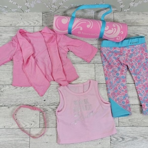 May include: A pink doll workout set including a jacket, tank top, leggings, headband, and a rolled yoga mat with a blue strap. The leggings have a pink and blue pattern. The tank top has silver text.