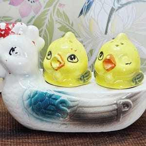 May include: A vintage ceramic salt and pepper shaker set featuring a white hen with a red comb and two yellow chick shakers. The hen has blue wing accents and the chicks have black eyes and orange beaks. The set is designed to sit on a table.