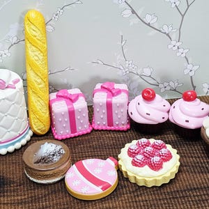 May include: A collection of miniature food items, including a white cake with a pink bow, a yellow baguette, pink gift boxes, cupcakes with cherries, and a tart with raspberries. The items are arranged on a brown surface.