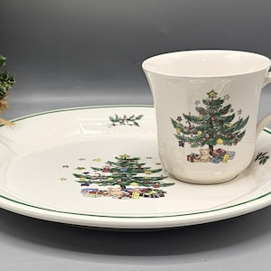 May include: A white ceramic Christmas tree-themed plate and cup set. The plate has a green rim and a green Christmas tree with presents and teddy bears. The cup has a green Christmas tree with ornaments and a green rim.
