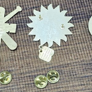 May include: A selection of metal pin badges and their backs. The badges include a sunburst design, a figure with arms outstretched, and a circular badge. The backs have pin clasps for secure attachment.