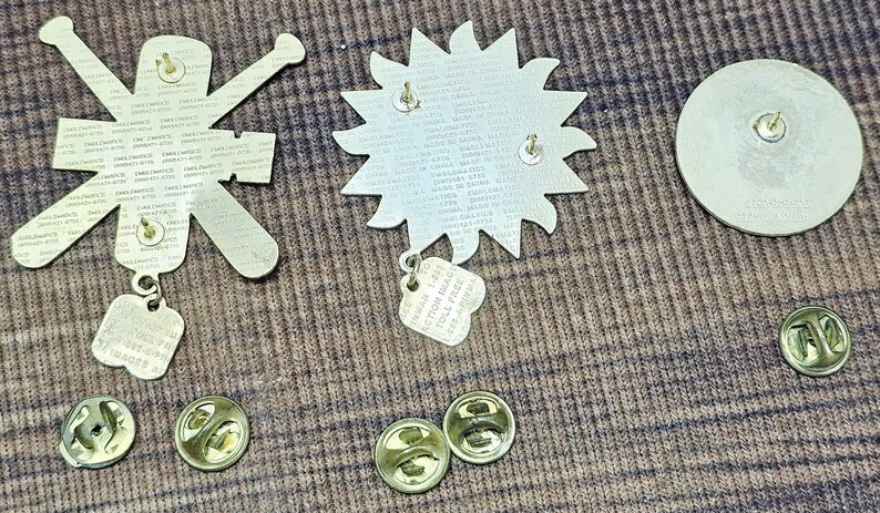 May include: A selection of metal pin badges and their backs. The badges include a sunburst design, a figure with arms outstretched, and a circular badge. The backs have pin clasps for secure attachment.