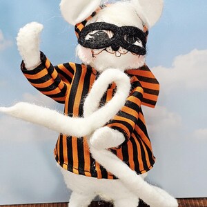 May include: A white mouse figurine wearing a black mask and an orange and black striped outfit. The mouse is holding a long, white, curved tail. The background is a blue sky with white clouds.