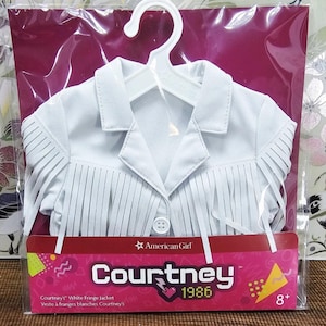 May include: White fringe jacket for a doll, displayed on a white hanger inside a clear plastic package. The jacket has a collared neckline and button closure. The packaging includes the text "American Girl" and "Courtney 1986".