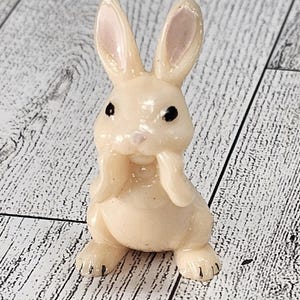 May include: A small, cream-colored bunny figurine with black eyes and pink-lined ears. The bunny is in a seated position, with its paws near its face. The figurine has a glossy finish and is set against a light wood-grain background.