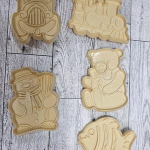 May include: Five beige plastic cookie cutters in various shapes: a train, a car, a snowman, a teddy bear, and a fish. The cutters are arranged on a light wood-grain surface.